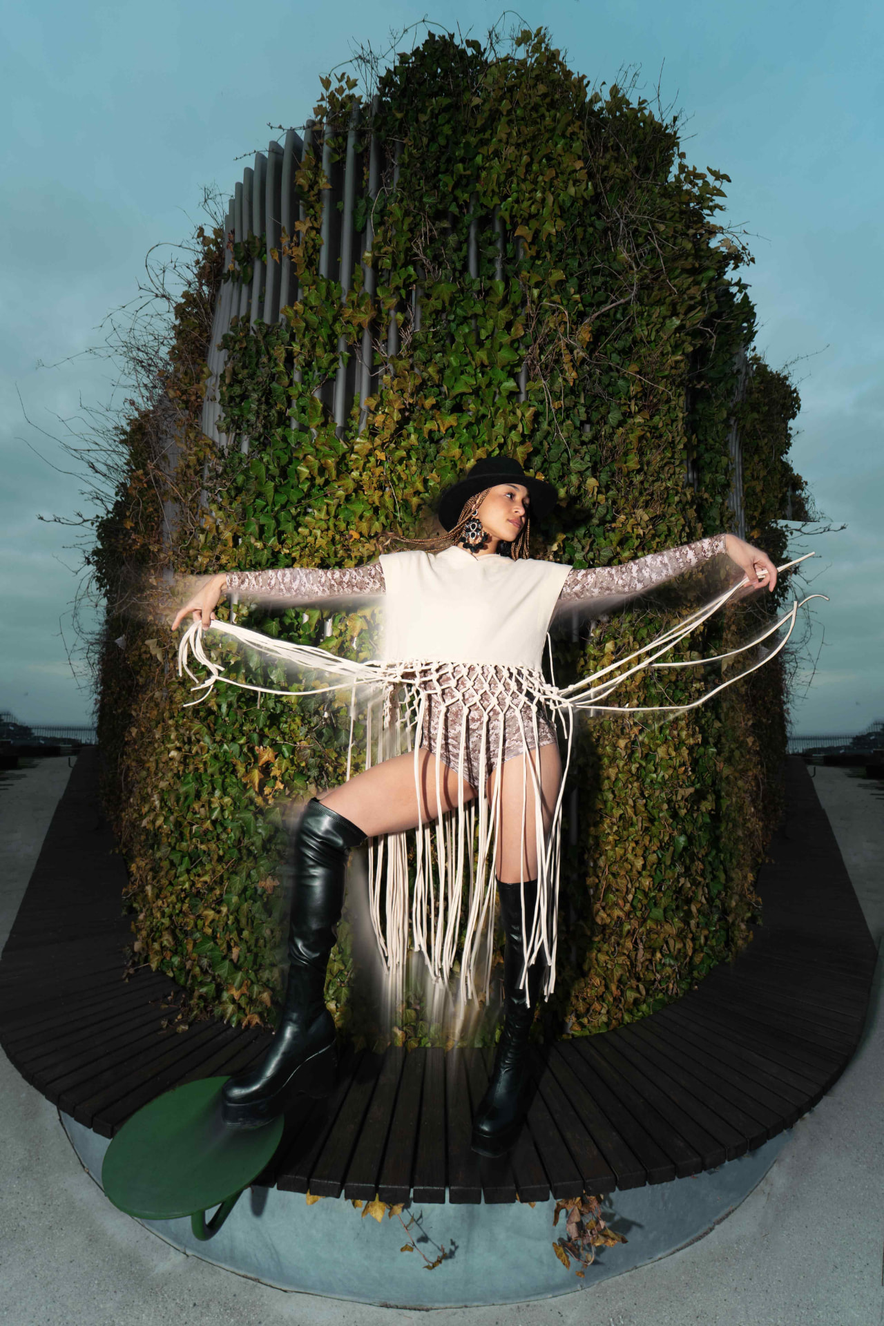 Noreena McCleave wearing a wide-brim hat and flowing fringed outfit, posed against a circular ivy-covered structure at dusk