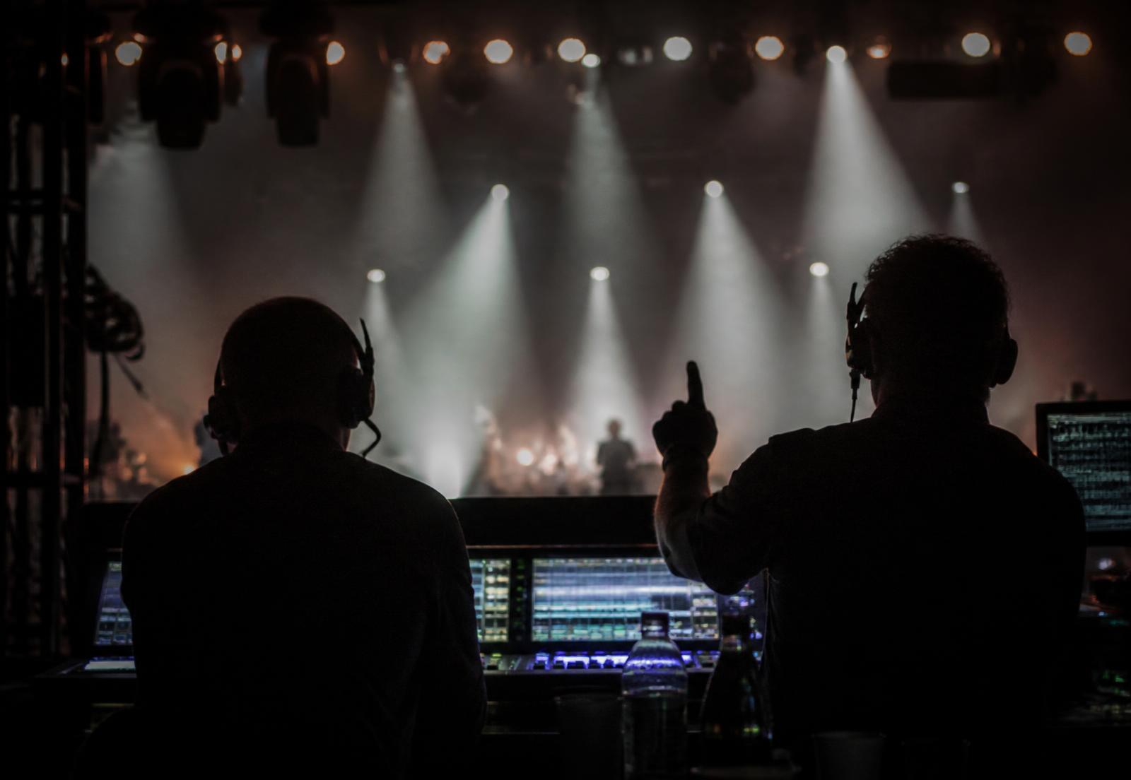 Crew in headsets working a lighting desk with stage beams and haze behind them